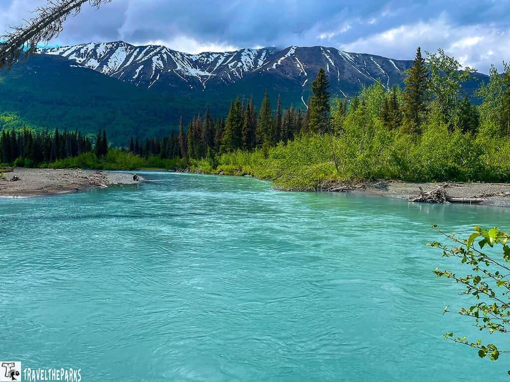On the road to Homer, a mountain landscape with a turquoise river, flanked by green trees and snowy peaks in the background.