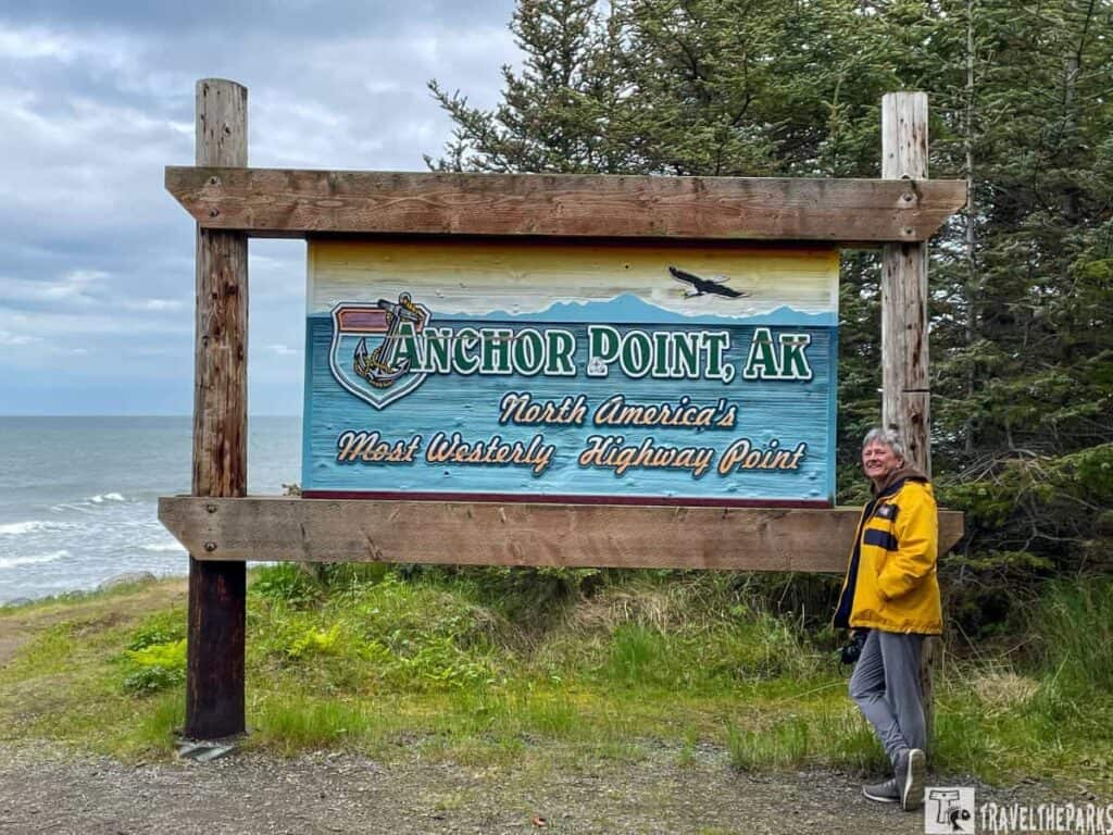 A sign for Anchor Point, AK, stating it is North America's Most Westerly Highway Point, with ocean and trees in the background.