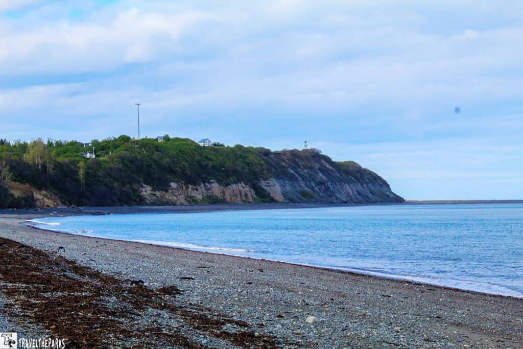 A pebbled beach next to calm blue water, with green cliffs and structures in the background under a cloudy sky.