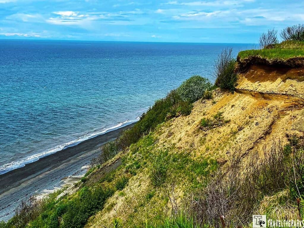 An Alaskan coastal landscape with a prominent cliff in the foreground sloping down toward a beach. The cliff is composed of layers of tan and brown earth, dotted with patches of green vegetation and low shrubs. Below the cliff lies a narrow strip of dark sandy beach extending along the shoreline. The expansive ocean fills the middle and background, showcasing varying shades of blue as it stretches to the horizon. The sky above is mostly covered with clouds, offering glimpses of blue. The overall scene gives a sense of tranquility and natural beauty.