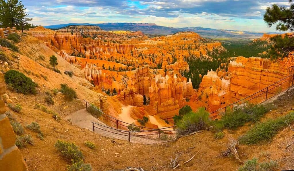 Panoramic view of Bryce Canyon National Park, featuring orange rock spires (hoodoos), a fence in the foreground, and a cloudy sky overhead.