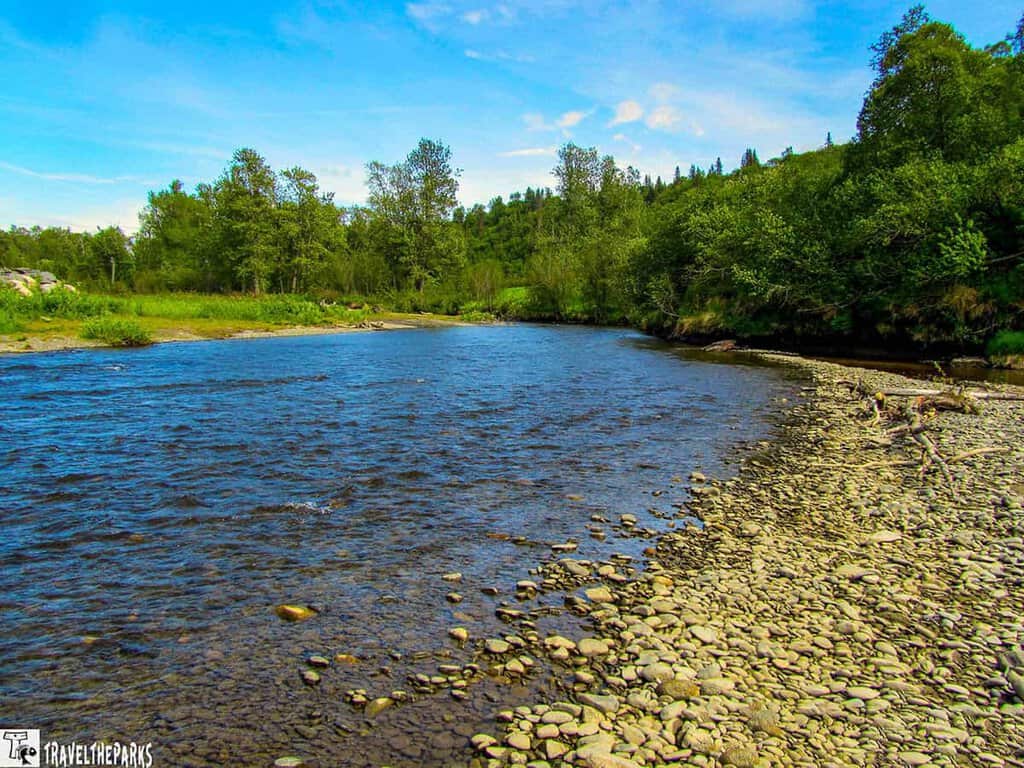 A peaceful Deep River scene with a pebbly shoreline and lush greenery under a blue sky.