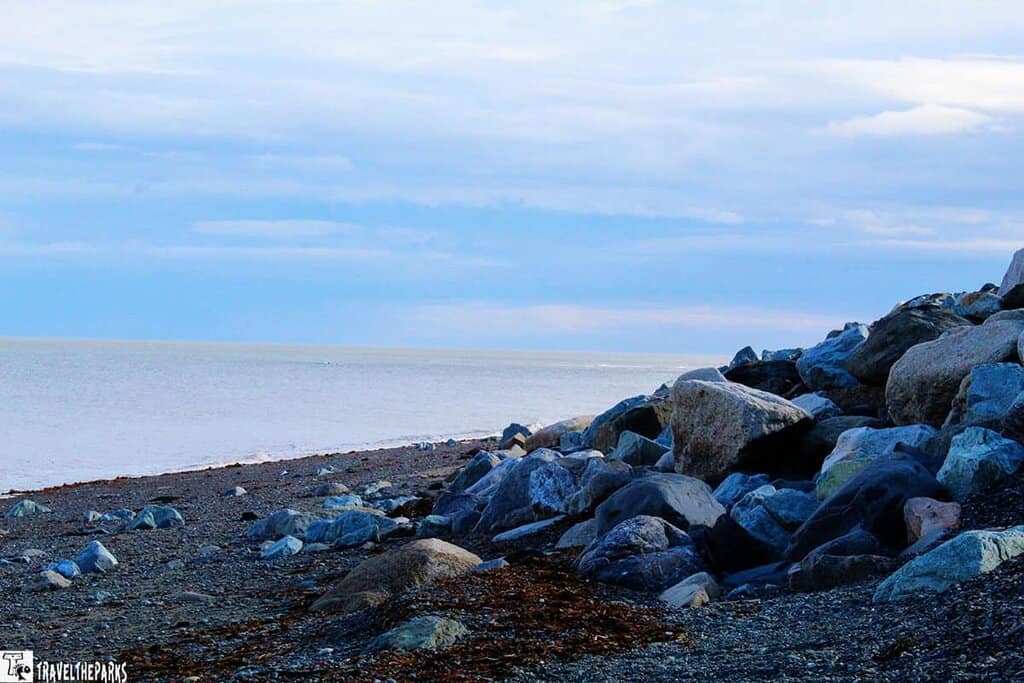 Rocky shoreline with large boulders and a gravel beach meeting a calm body of water under a cloudy sky.