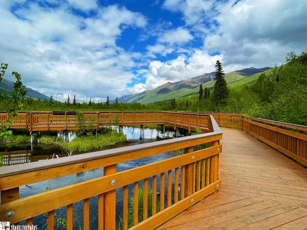 Wooden boardwalk at Eagle River Nature Center with mountains and cloudy blue sky in the background.

