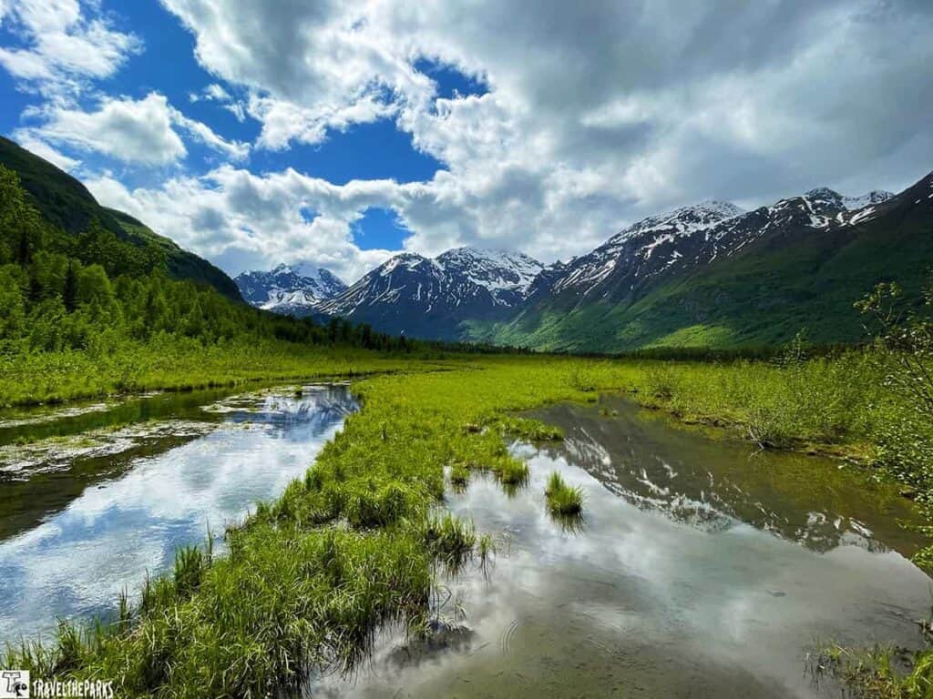 A tranquil landscape at Eagle River Nature Center with a reflective stream, green fields, and snow-capped mountains under a partly cloudy sky.

