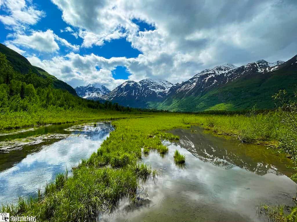 A tranquil landscape at Eagle River Nature Center with a reflective stream, green fields, and snow-capped mountains under a partly cloudy sky.