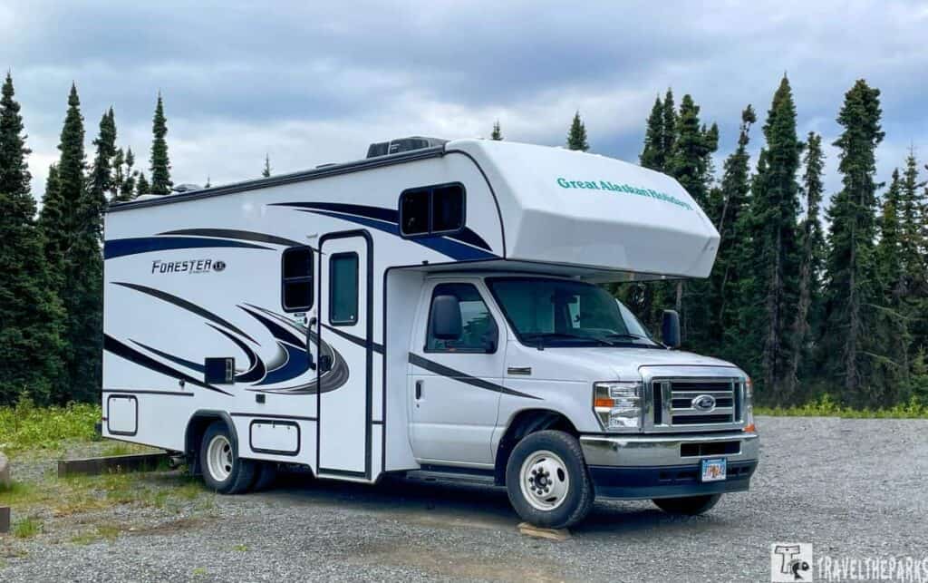 a white recreational vehicle (RV) parked on a gravel area, surrounded by a backdrop of tall evergreen trees under an overcast sky. The RV is marked with swooping blue and black decals on its side and features the model name "Forester" with a logo. The front section of the RV is a truck cab with a white exterior and silver grille, and it has a door and windows. There is also an over-cab sleeping area, which is typical for such camper vans. The side of the RV displays a large window and a side mirror. The background includes dense trees, indicating a forested location.