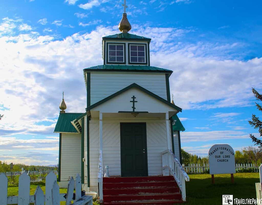 A small white Holy Transfiguration of Our Lord Russian Orthodox Church with green roofs and an Orthodox cross, set against a partly cloudy blue sky.