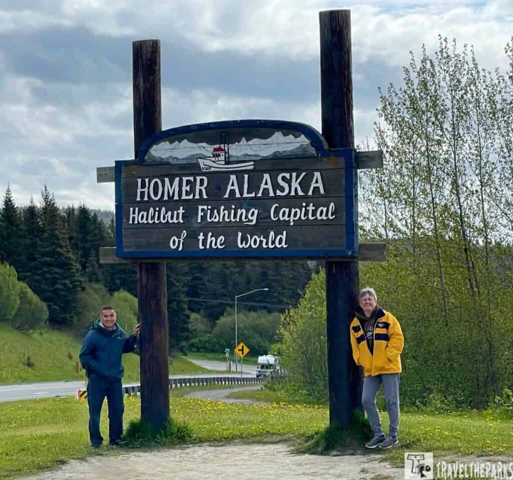 A sign reading "Homer Alaska, Halibut Fishing Capital of the World" with two people standing beside it on a scenic roadside.