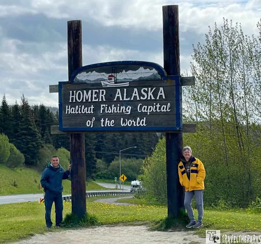 A sign reading "Homer Alaska, Halibut Fishing Capital of the World" with two people standing beside it on a scenic roadside.