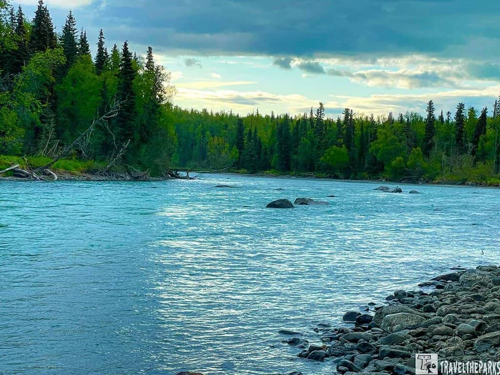 Kasilof river with clear blue water flows alongside a rocky shore, bordered by dense green trees, under a partly cloudy sky.