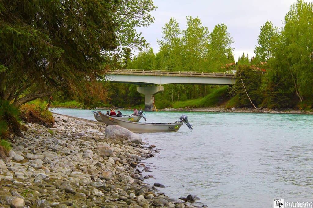 A serene Kasilof river scene in an area likely along the road to Homer, Alaska. In the foreground, a rocky shoreline lines the left side of the image, with smooth, round stones varying in shades of gray. Adjacent to the shoreline, two small motorboats are anchored near the water's edge. Several people are seated in the boats, wearing outdoor clothing suitable for the temperate climate. The river is a vivid turquoise color, reflecting the surrounding greenery and sky. In the background, a concrete bridge spans the river, connecting lush, forested banks. The area is densely populated with tall, green trees that frame the entire scene, their foliage bright against the overcast sky.
