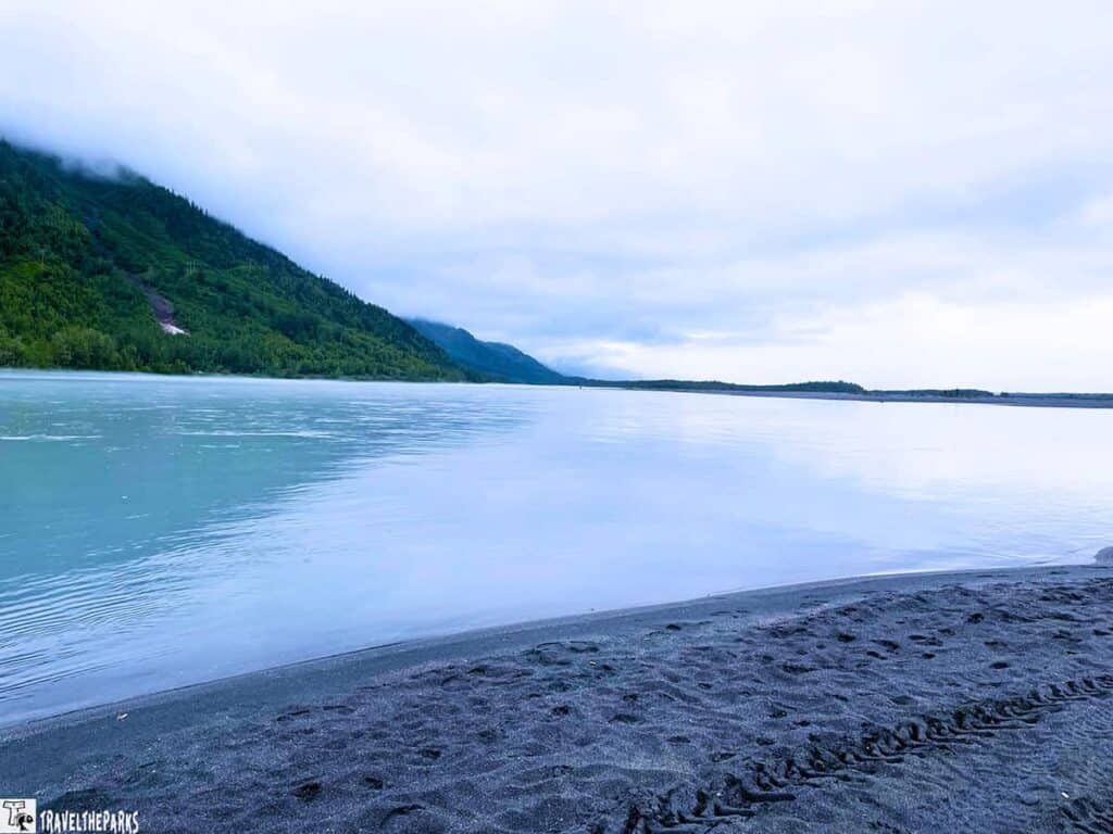 Serene view of the Knik River with a sandy shoreline and forested mountains in the background under cloud-covered skies.

