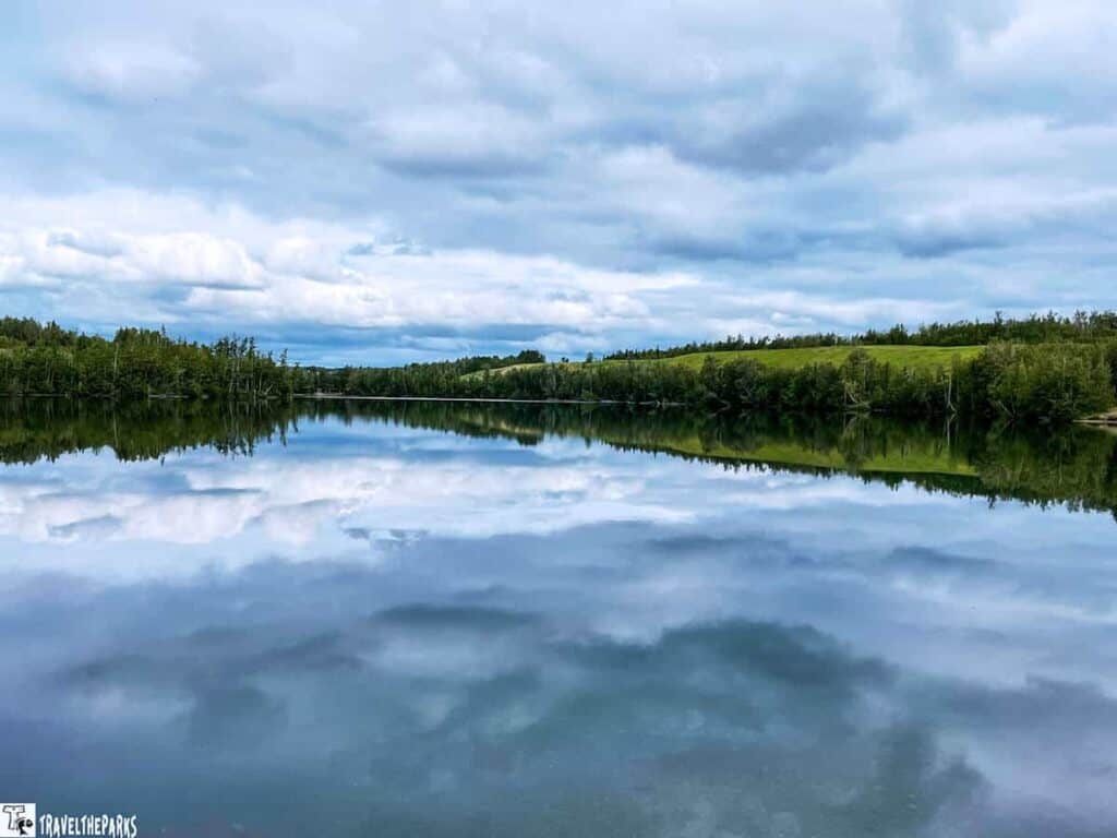 A peaceful lake reflecting the cloudy sky and surrounding green trees at Matanuska Lakes State Recreation Area.

