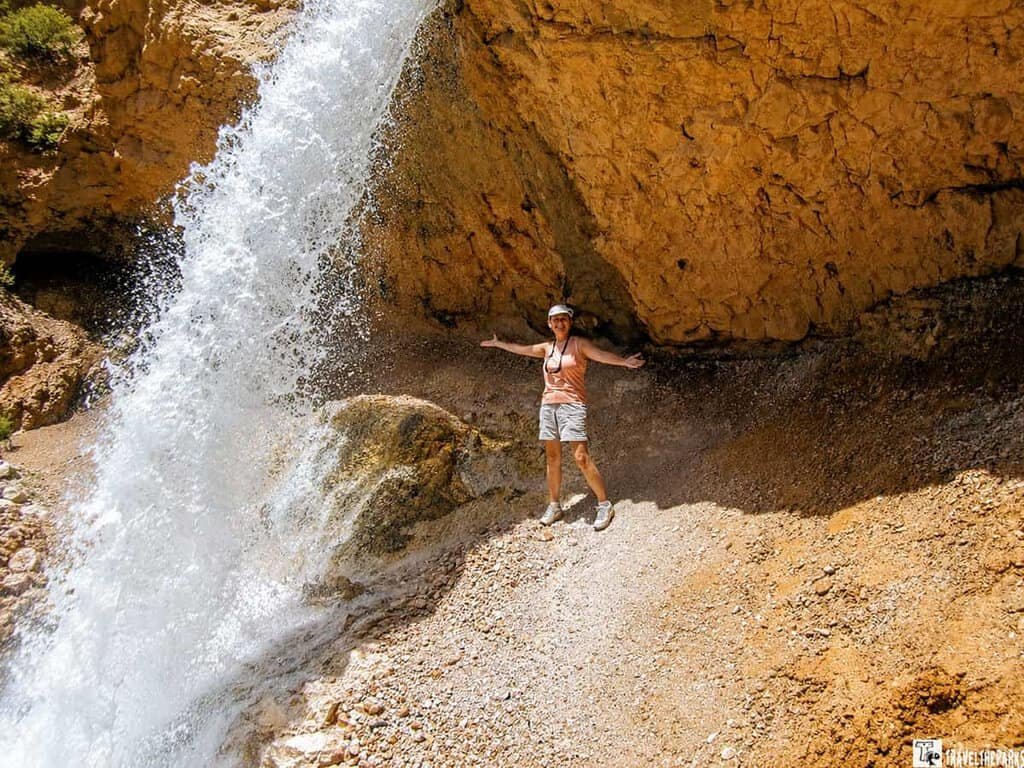 A woman stands in front of a cascading waterfall at Mossy Cave Loop in Bryce Canyon, with her arms outstretched. The rocky cliff behind her features textured orange-brown stone.