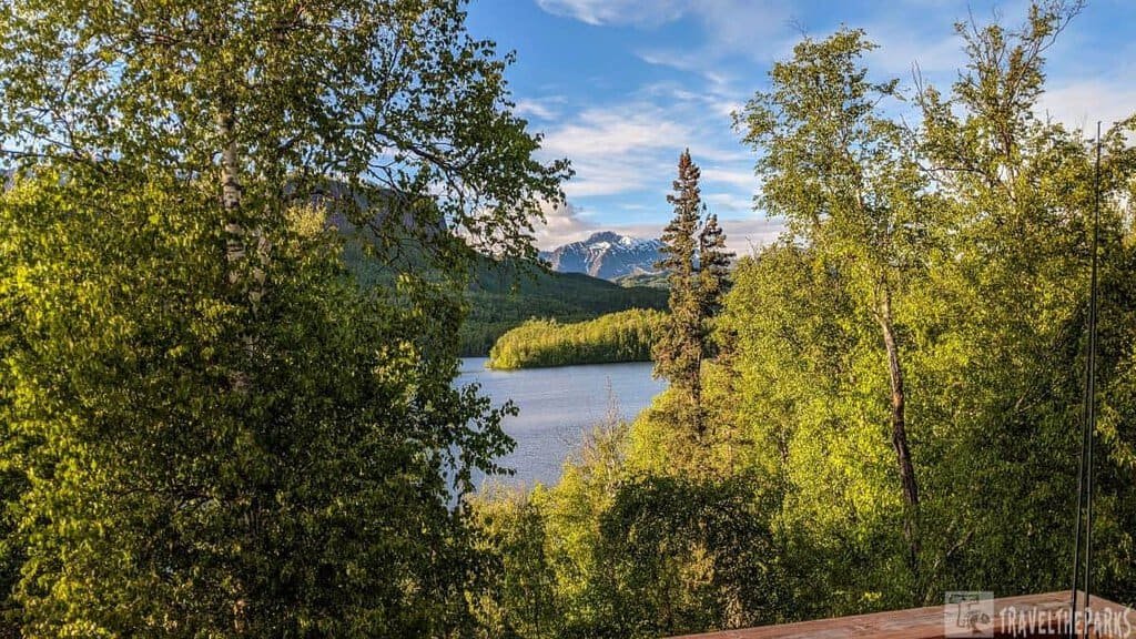 A scenic view of a lake surrounded by dense green forest and snow-capped mountains under a blue sky.