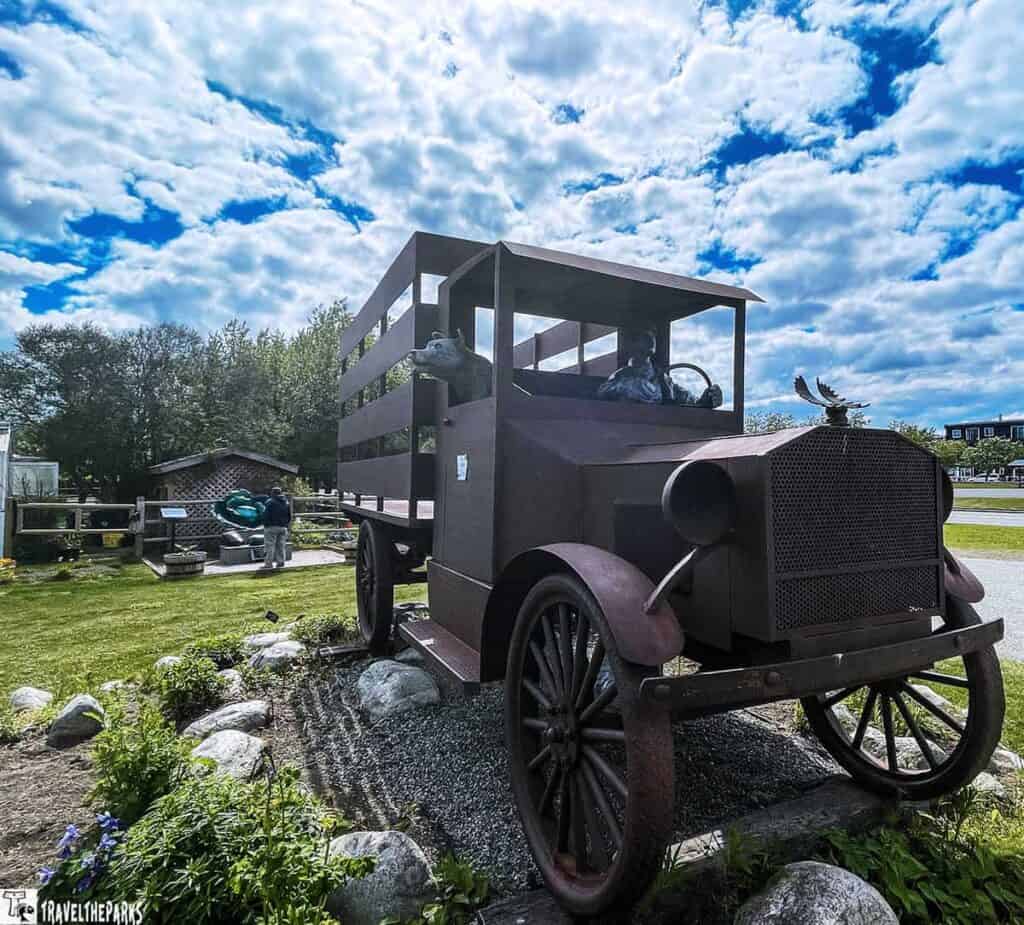 A large rusty sculpture of a vintage truck with animal statues inside, set in an outdoor display on a sunny day with cloudy skies.