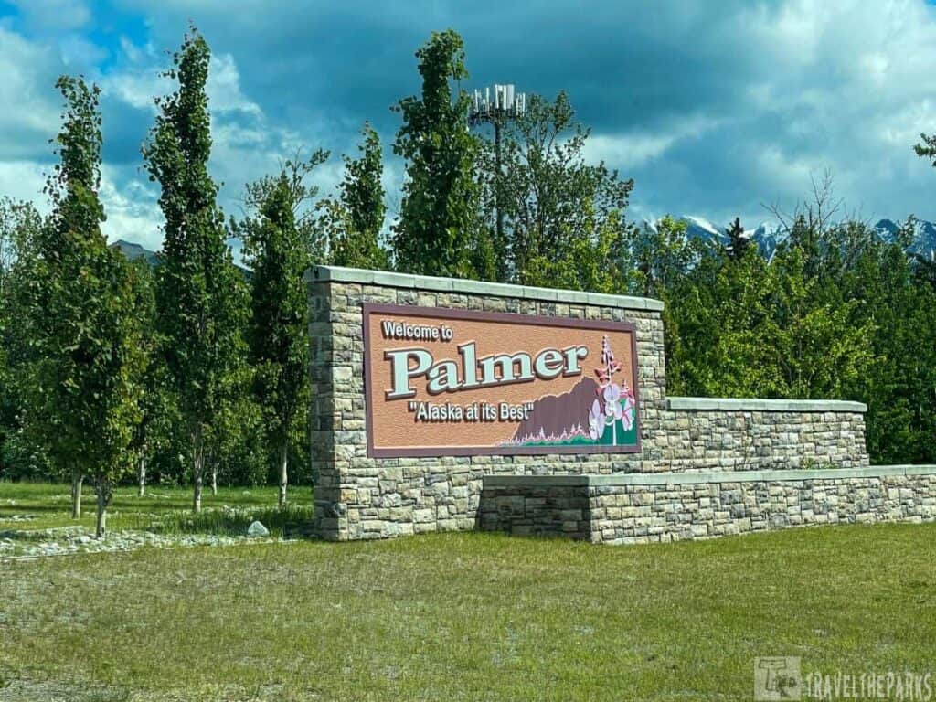A stone sign reads "Welcome to Palmer, 'Alaska at its Best'" surrounded by trees and a partly cloudy sky.


