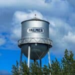 A silver water tower with "PALMER" written on it against a cloudy sky and surrounded by tree foliage.
