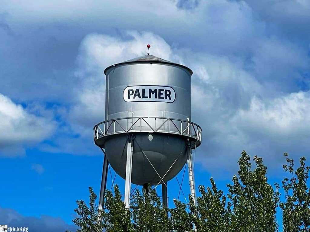 A silver water tower with "PALMER" written on it against a cloudy sky and surrounded by tree foliage.