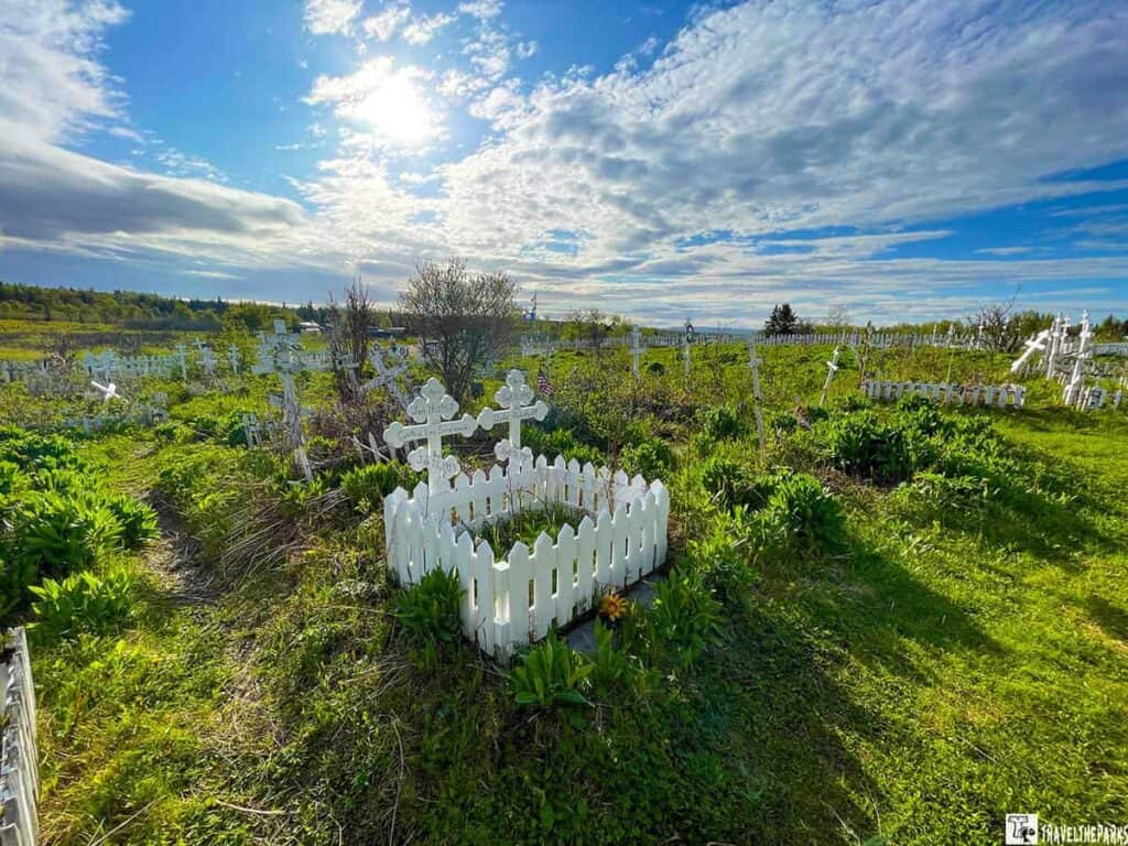 A rural cemetery with white crosses and picket fences under a bright, partly cloudy sky.