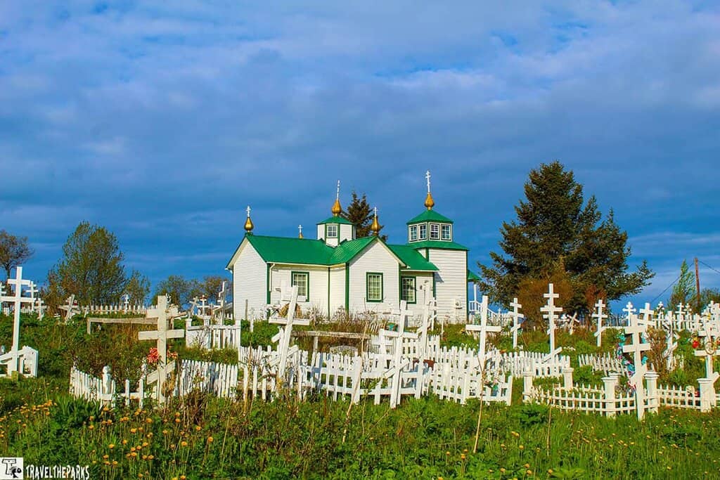 A white Holy Transfiguration of our Lord Russian Orthodox Church with a green roof and golden domes surrounded by a cemetery with white crosses and fences.