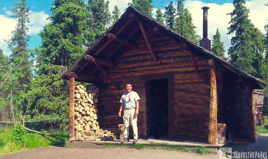 A log cabin in a forest with a person standing at the doorway and stacked firewood nearby.