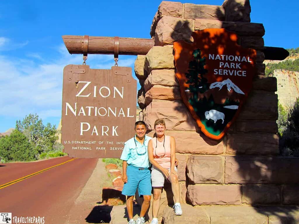 Entrance sign to Zion National Park with visitors standing beside the National Park Service emblem.