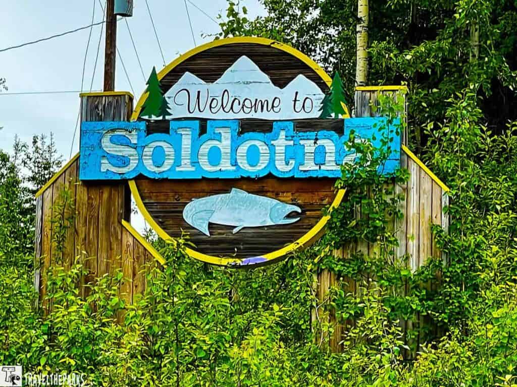 Welcome sign for Soldotna with mountains, pine trees, and a fish illustration surrounded by greenery.