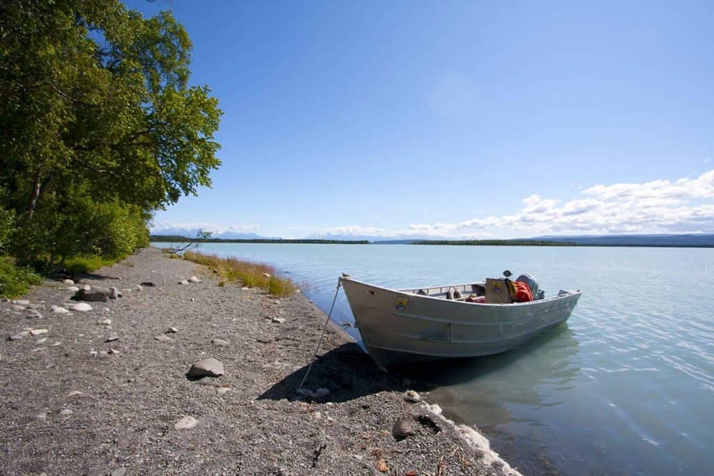 A small boat moored on a pebble shore beside a calm lake Tustumena with trees and mountains in the distance under a blue sky.