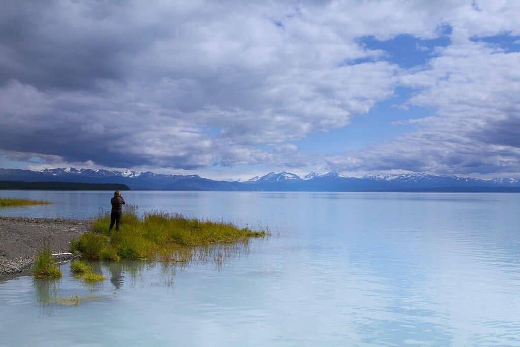 a tranquil scene of a person standing on a narrow strip of land that juts into a large, calm body of Tustumena Lake. The person, wearing dark clothing, appears to be photographing or observing the scenery. The foreground includes patches of lush green grass and gravel. The expansive, mirror-like water reflects the blue and gray hues of the partly cloudy sky. In the background, a range of snow-capped mountains lines the horizon, adding depth to the scene. The sky above is a mix of bright blue with large, fluffy clouds, creating a dramatic and vast atmosphere.