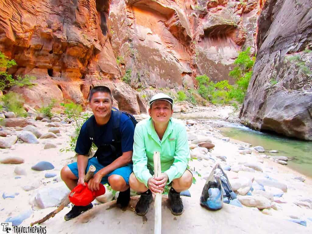 Two people kneeling on a sandy pathway in a narrow canyon with red rock walls and a stream.