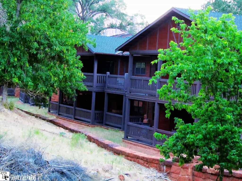 Zion Lodge-Watchman Building: Rustic wooden building with balconies against a backdrop of tall red sandstone cliffs and blue sky.