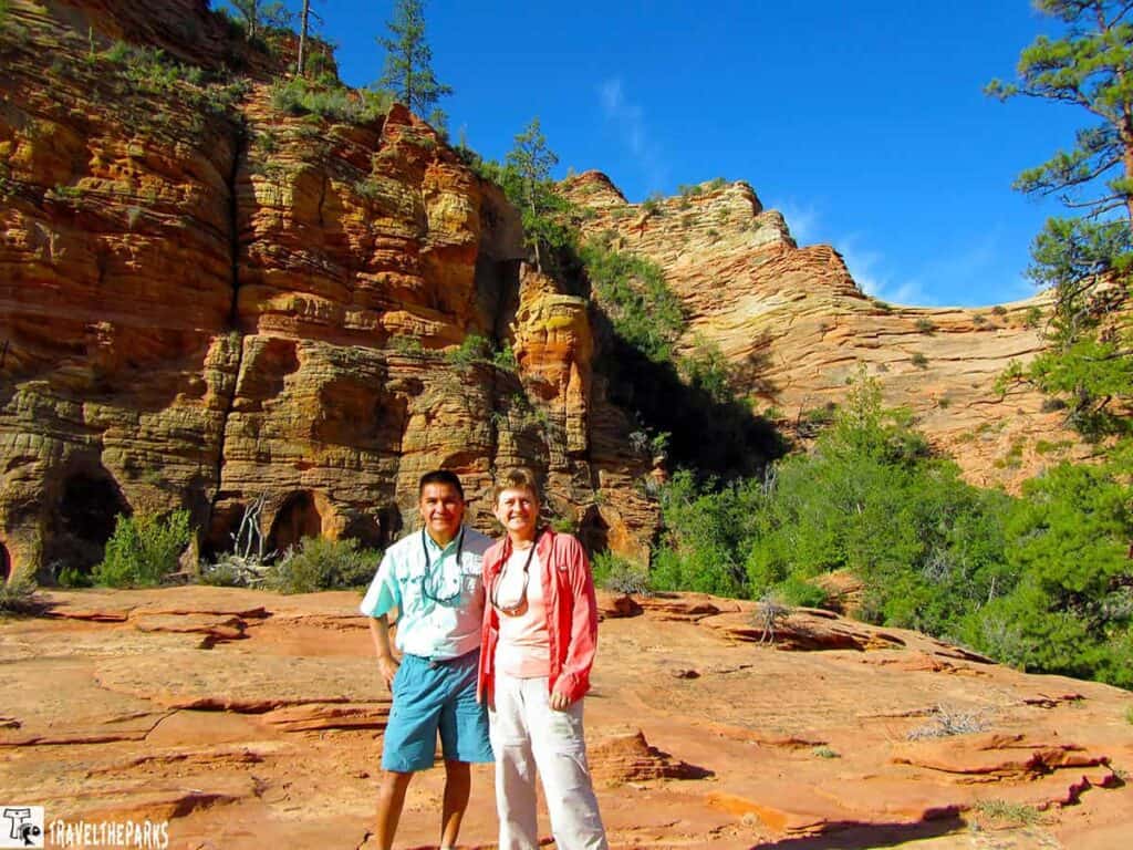 Two people standing on a rocky surface with red rock formations and a clear blue sky in the background.