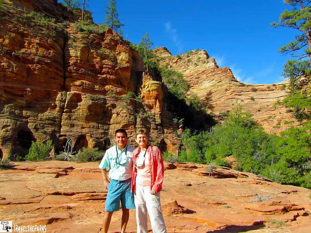 Two people stand on rock formations with layered sandstone cliffs behind them at Zion National Park.