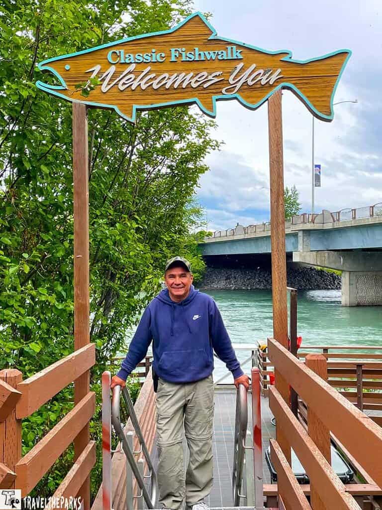 A man stands on a walkway under a fish-shaped sign that reads "Classic Fishwalk Welcomes You" next to a river.