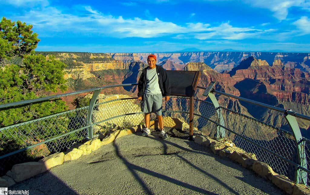 Man smiling at the Grand Canyon viewpoint. Vibrant blue sky, red canyon layers, and a railing surround him. The scene conveys awe and tranquility.