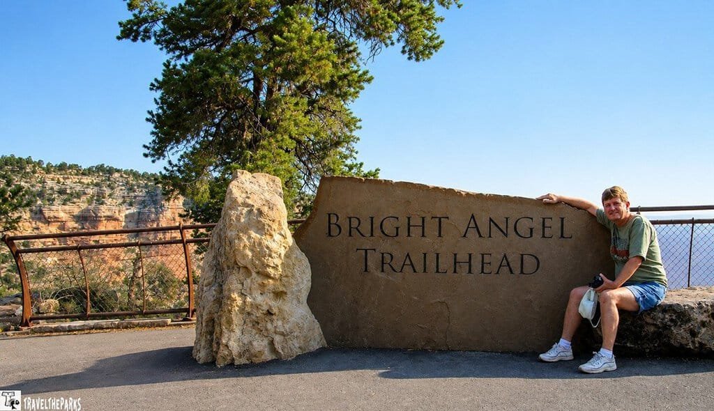 A person poses by the Bright Angel Trailhead sign at the Grand Canyon. The scene is sunny, with canyon views and a tree in the background, conveying adventure.