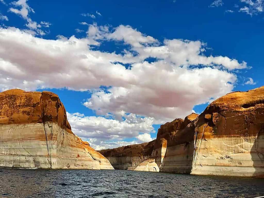 View of Lake Powell with layered sandstone canyon walls and a vibrant blue sky with clouds.

