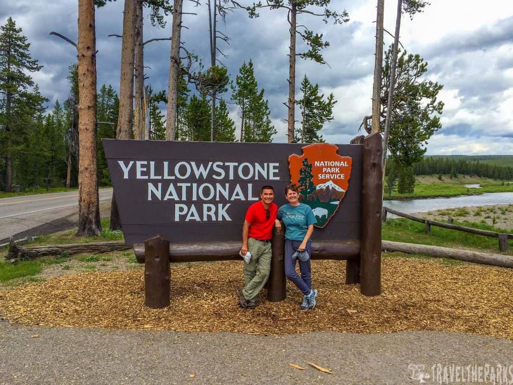 Yellowstone National Park Sign with two people stading in front. The snake river is in the background.