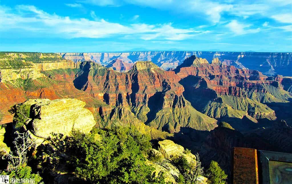 Deva-Brahma-and-Zoroaster-Temples: A vibrant view of the Grand Canyon under a clear blue sky. The canyon's layered rock formations reveal deep reds and browns, bordered by lush green trees.