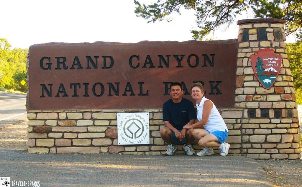 A smiling couple poses in front of the Grand Canyon National Park sign made of stone. Bright daylight enhances the scene's welcoming and adventurous feel.