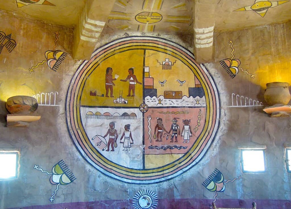 Native american-style mural with a colorful circular scene of people, animals, symbols, and boats, framed by hieroglyphic motifs on ochre walls.