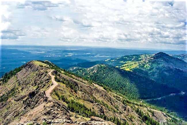 Panoramic view from Mt. Washburn, showing a winding trail, rocky terrain, and forested hills under a partly cloudy sky.