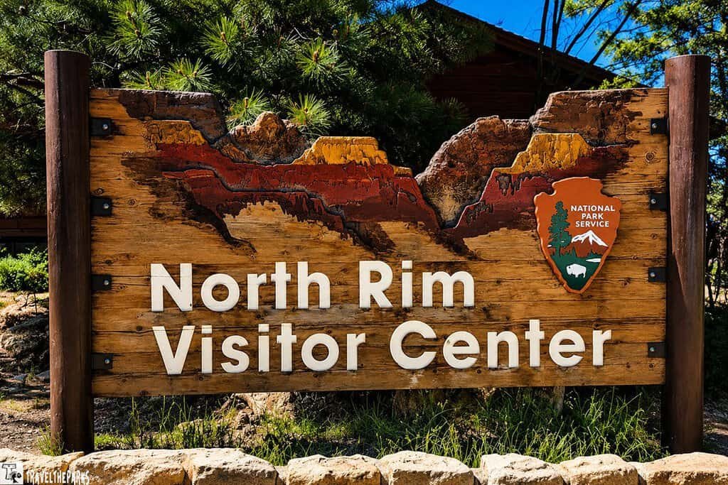 Wooden sign for "North Rim Visitor Center" with a landscape carving and the National Park Service logo. Surrounded by pine trees, conveying a welcoming, natural setting.