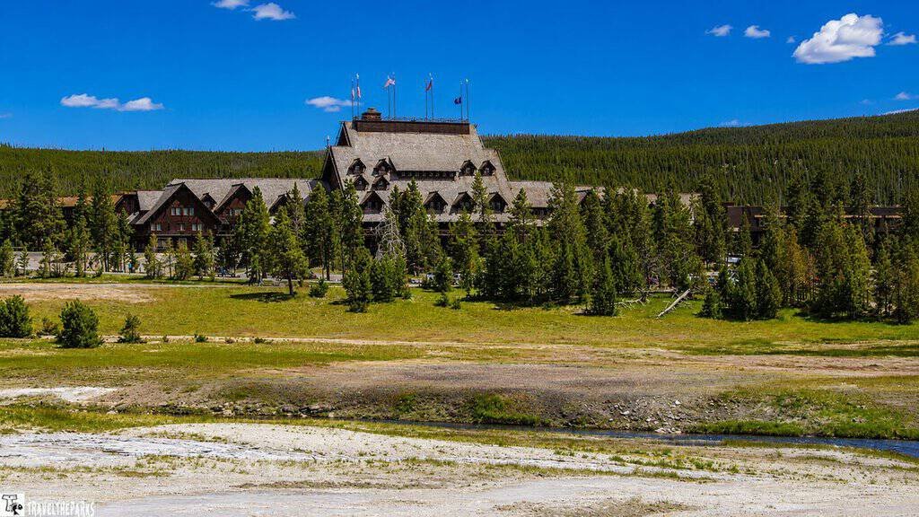 Old Faithful Inn-Yellowstone National Park: A large, rustic building with flags on the roof, surrounded by evergreen trees and grassy fields.