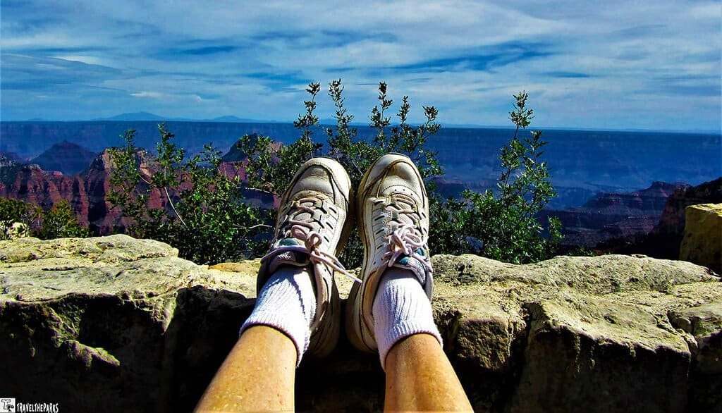 A pair of feet in white sneakers is perched on a ledge overlooking the expansive Grand Canyon. The view is vast, showcasing multicolored canyon layers.
