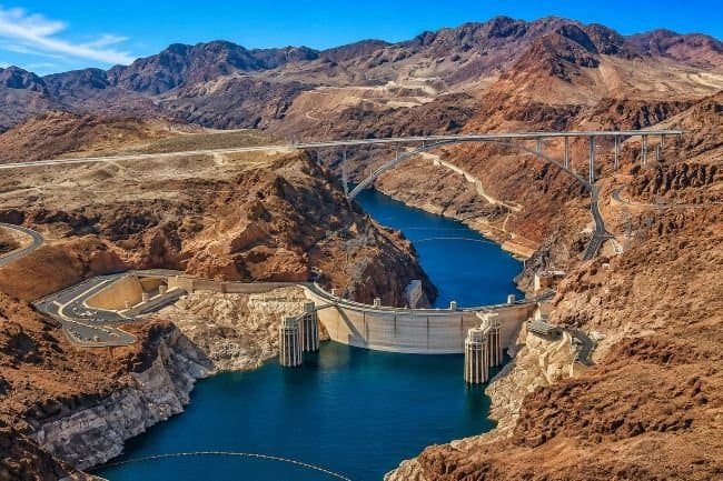 Helicopter View Over Hoover Dam: Aerial view of a dam with a reservoir between rocky cliffs and an overpassing bridge.