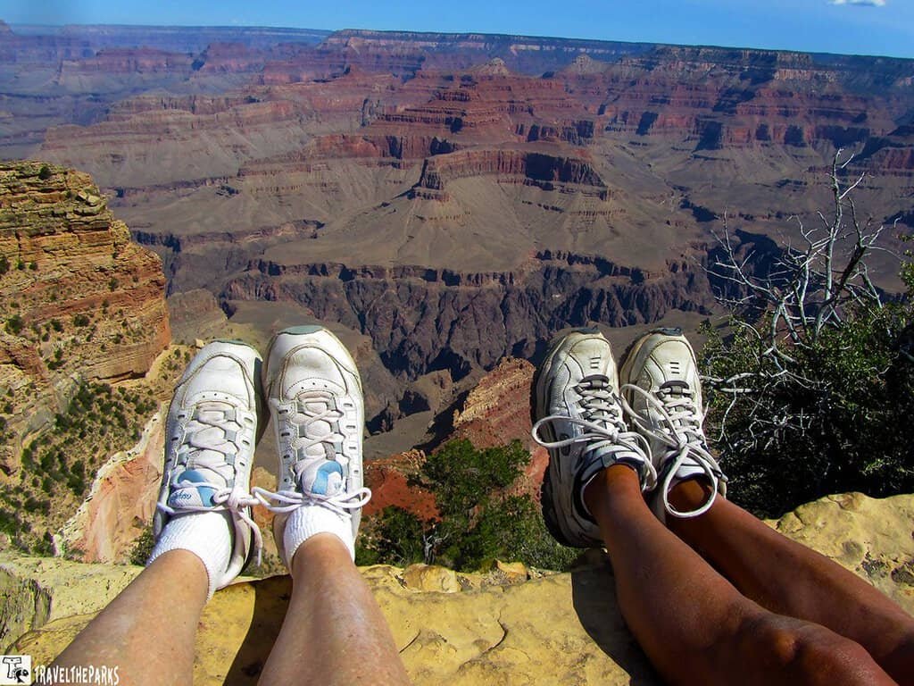 Two people wearing sneakers sit at the edge of the Grand Canyon, with legs extended over the vast, layered landscape under a clear blue sky.