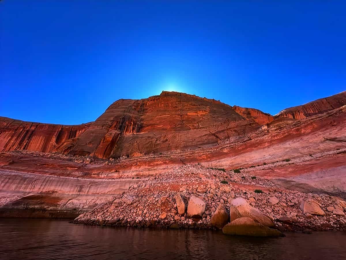 View of a sunlit sandstone cliff and Lake Powell under a bright blue sky.